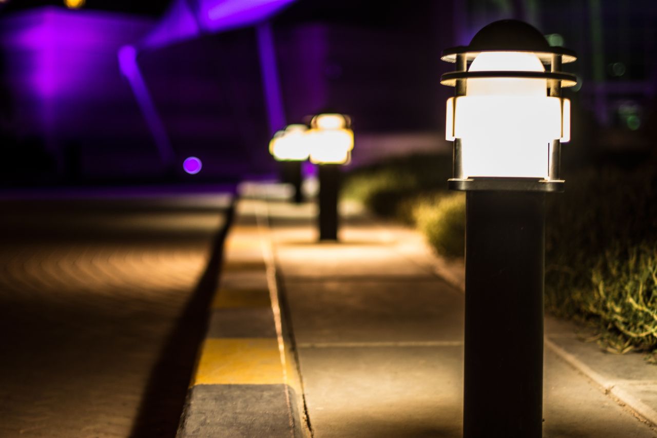 gallery-03 A scenic night view of illuminated lamp posts lining a paved path with a bokeh effect.