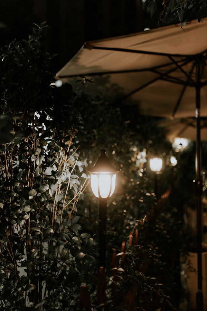 services-01 Illuminated garden path with lanterns, umbrellas, and shrubs creating a tranquil nighttime atmosphere.